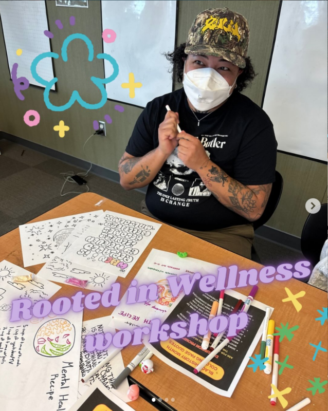 Person wearing a camouflage cap and face mask sits at a table coloring printed wellness worksheets during a “Rooted in Wellness” workshop, with markers and activity pages spread out.